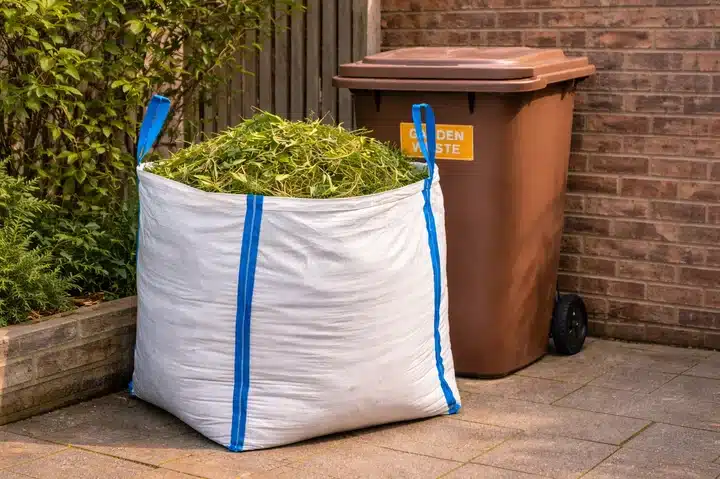 Garden waste bulk bag filled with green cuttings beside wheelie bin.