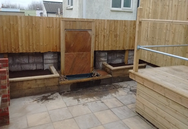 Contemporary patio with timber fencing, raised planters, and a slate water feature.