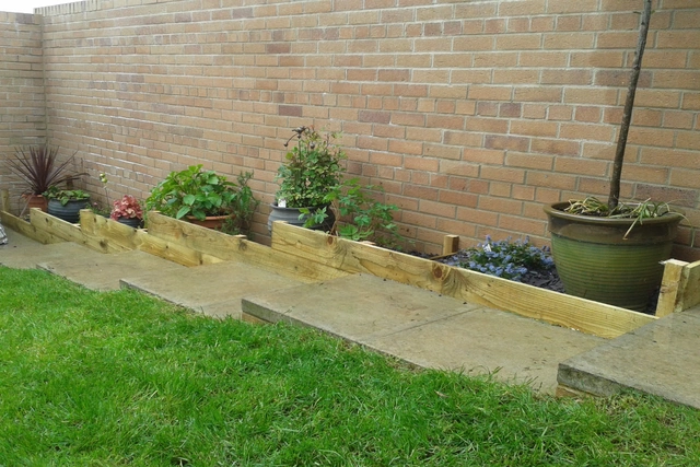 Timber-edged garden border with planted pots and flowers alongside a lawn and patio path.